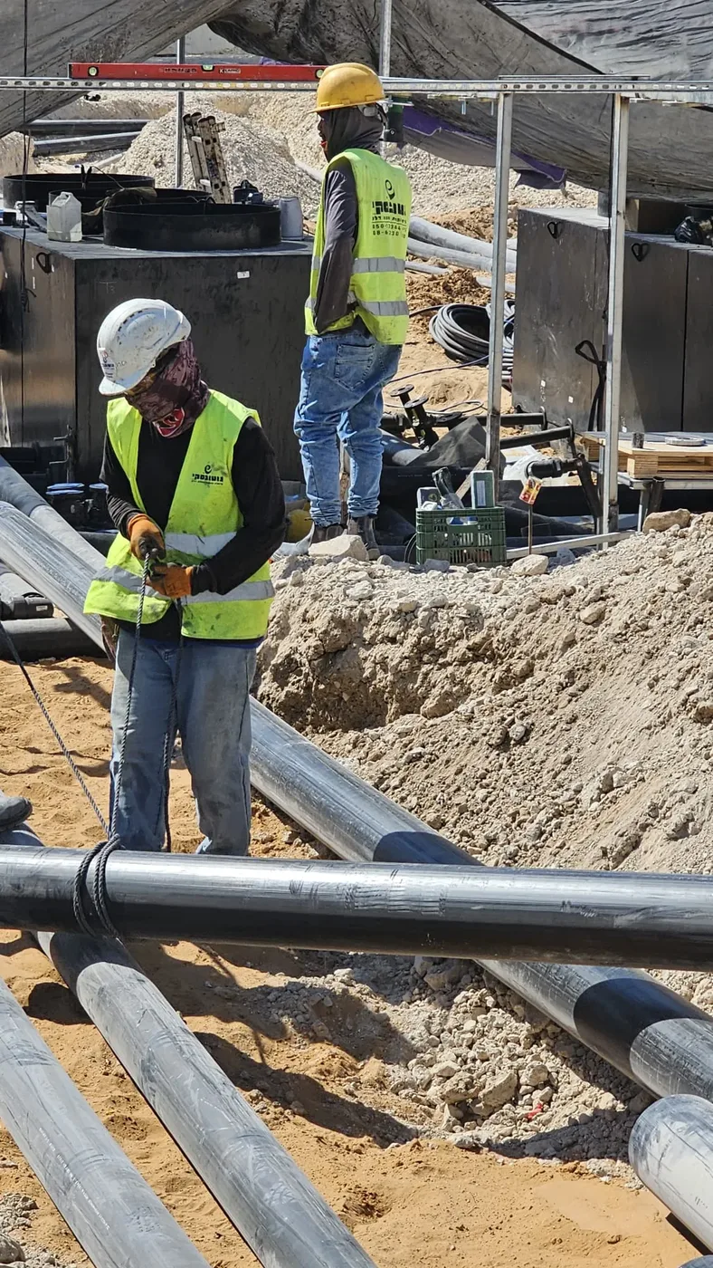 שני עובדים באפודים זוהרים מניחים צנרת דלק, מיכלים ברקע