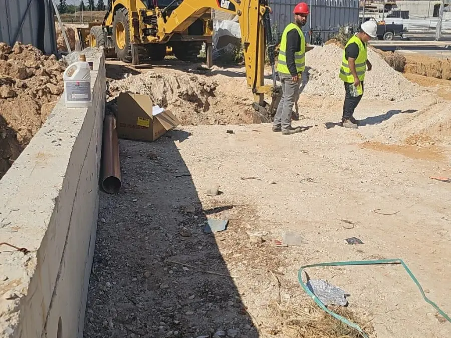 מחפרון קטרפילר ושני עובדים באפודים בחפירה לצד קיר תומך