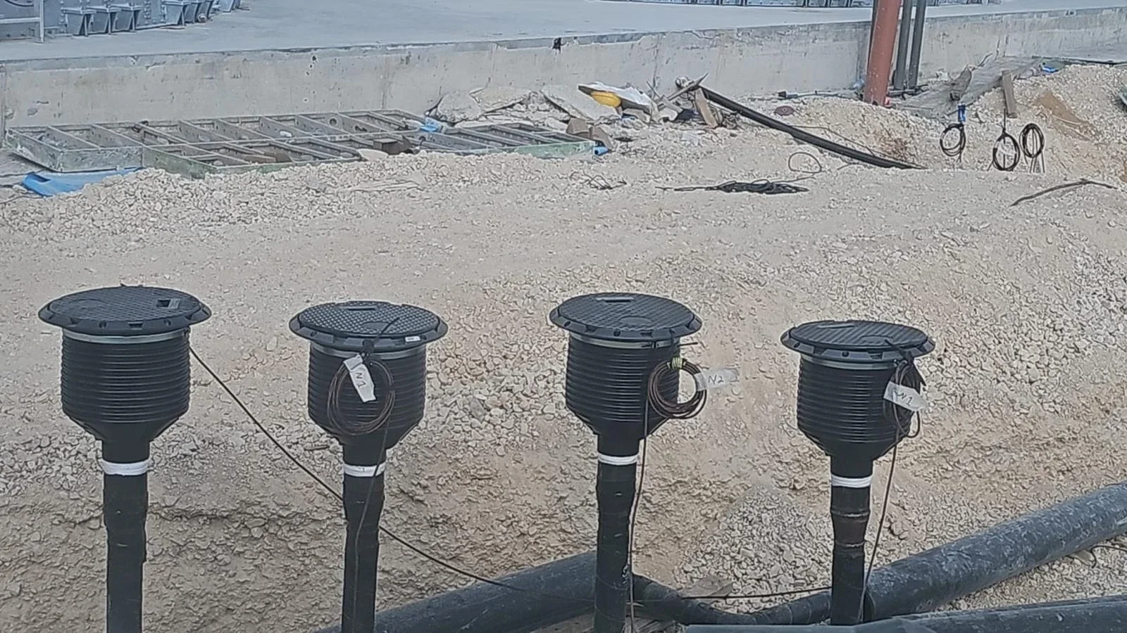 ארבע שוחות פלסטיק שחורות עגולות עם כבלים מסומנים, על חול ליד קיר בטון