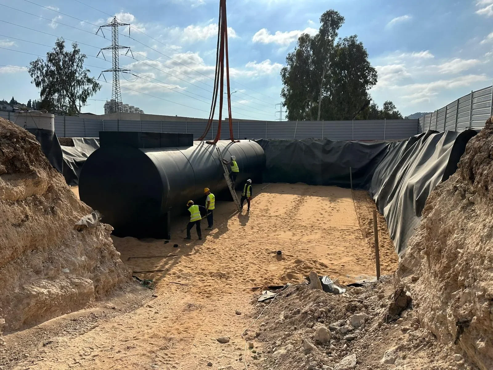 מיכל דלק שחור מונח בבור על חול, ארבעה עובדים סביבו, סולם נשען עליו, רתמות המנוף מעל