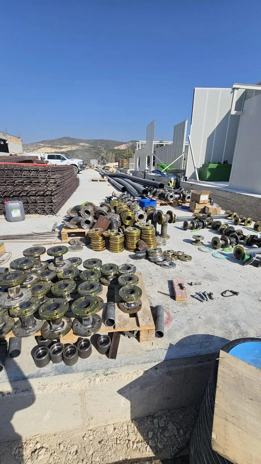 ערימות של אוגני פלדה, ספחים ואביזרי צנרת מסודרים על משטח בטון, צינורות ומבנה ברקע