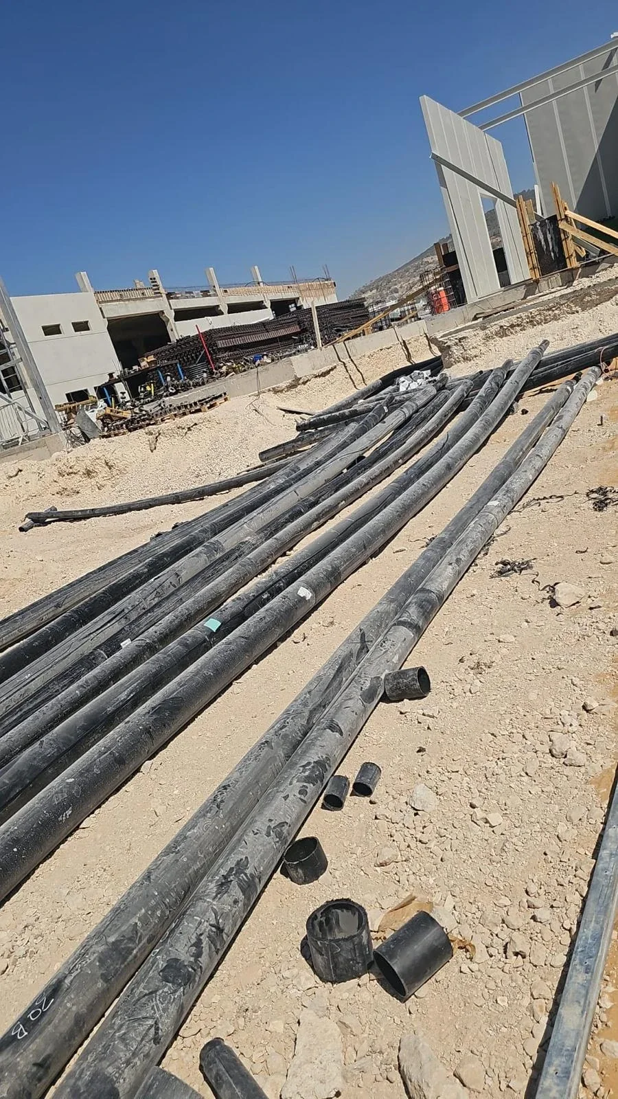 צינורות שחורים ארוכים מונחים על רחבת חצץ, מבנה בבנייה ברקע