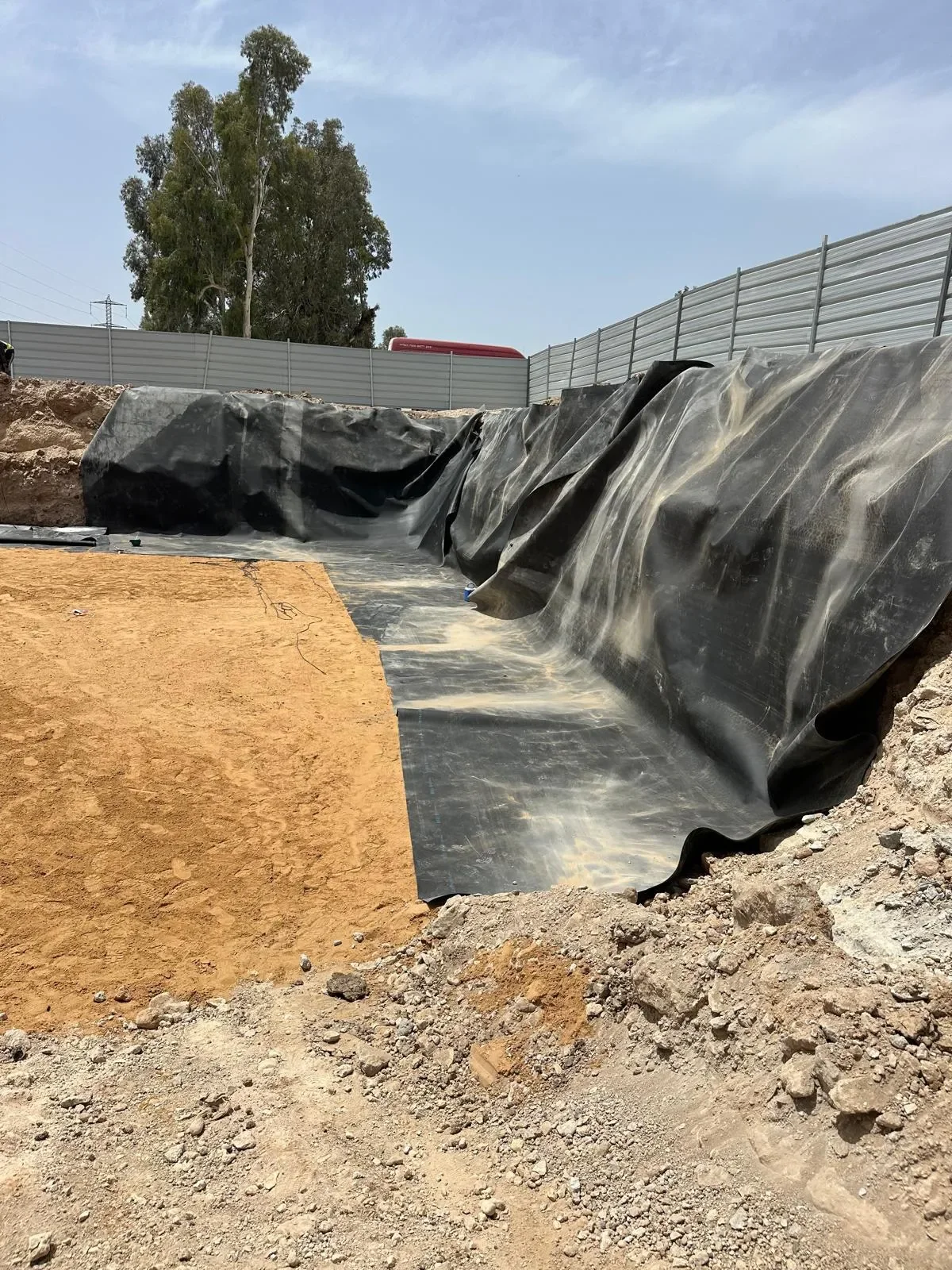 בור חפור עם יריעת איטום שחורה על הדפנות ושכבת חול בהירה בתחתית, גדר אתר ועץ אקליפטוס ברקע