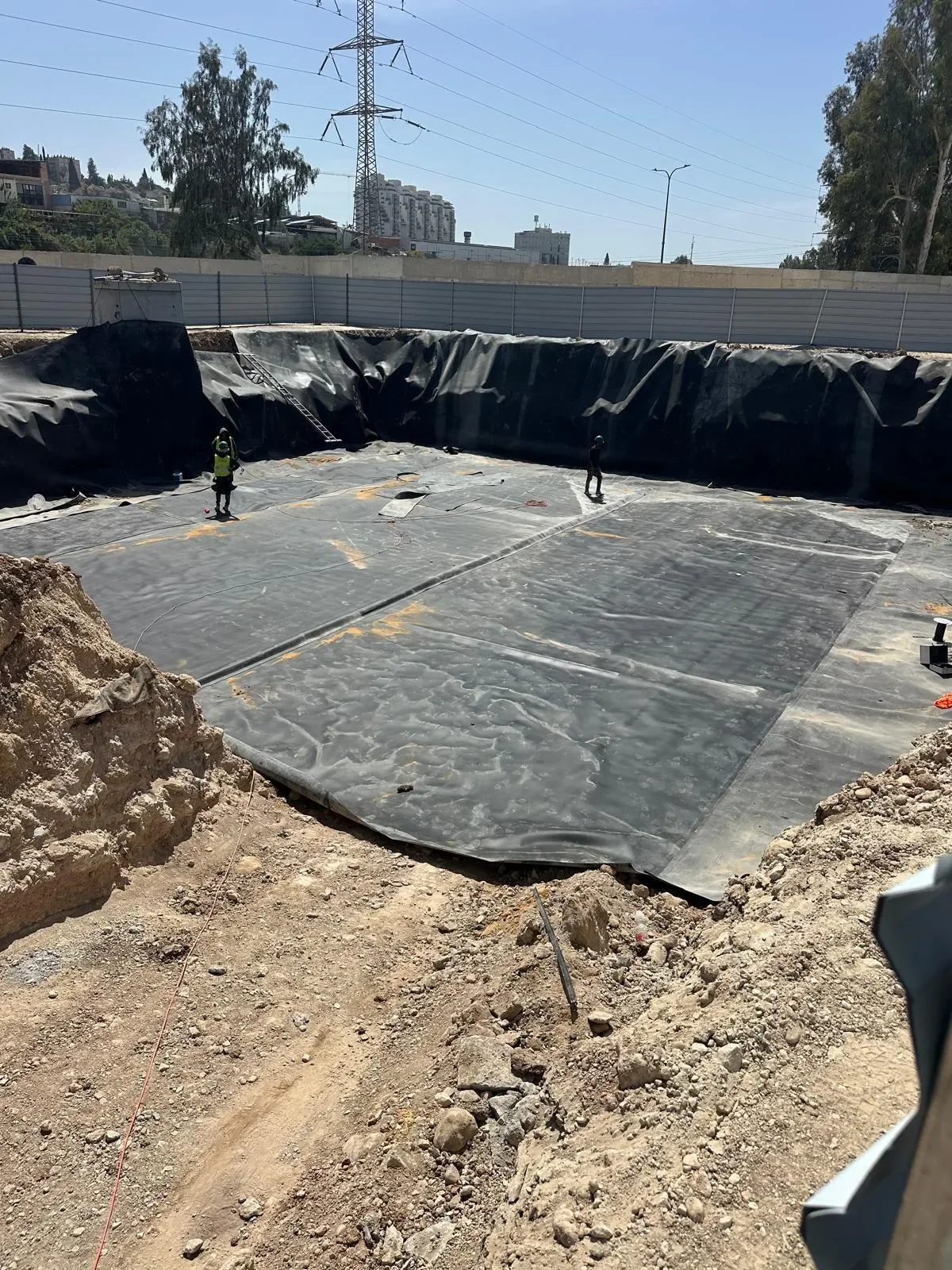 מבט רחב על בור מרובע גדול מכוסה יריעה שחורה, שני עובדים בבור, גדר אתר ובניינים ברקע