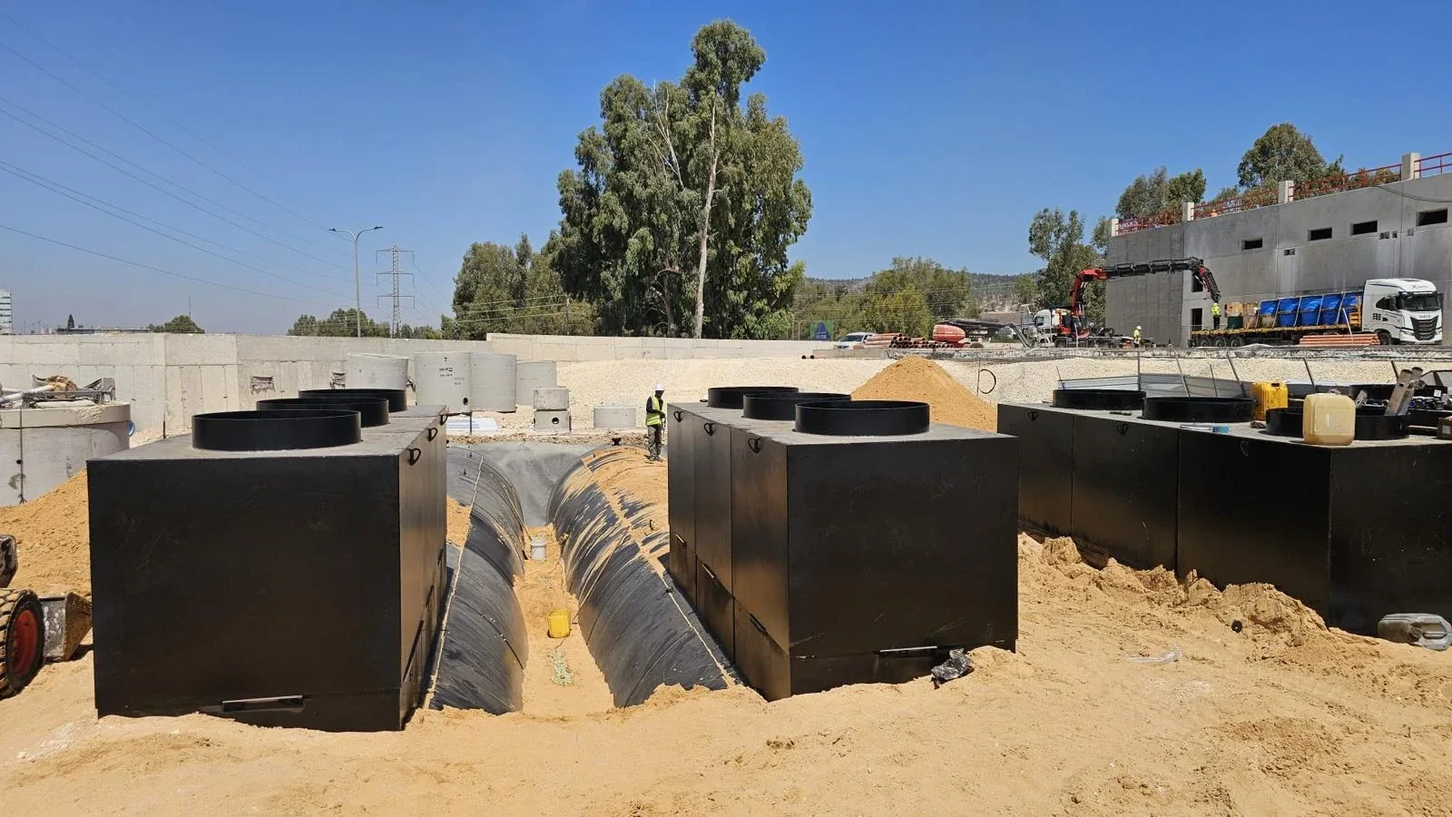 שלושה תאי שוחה שחורים מרובעים בולטים מעל חול, מבנה בבנייה וגדר ברקע