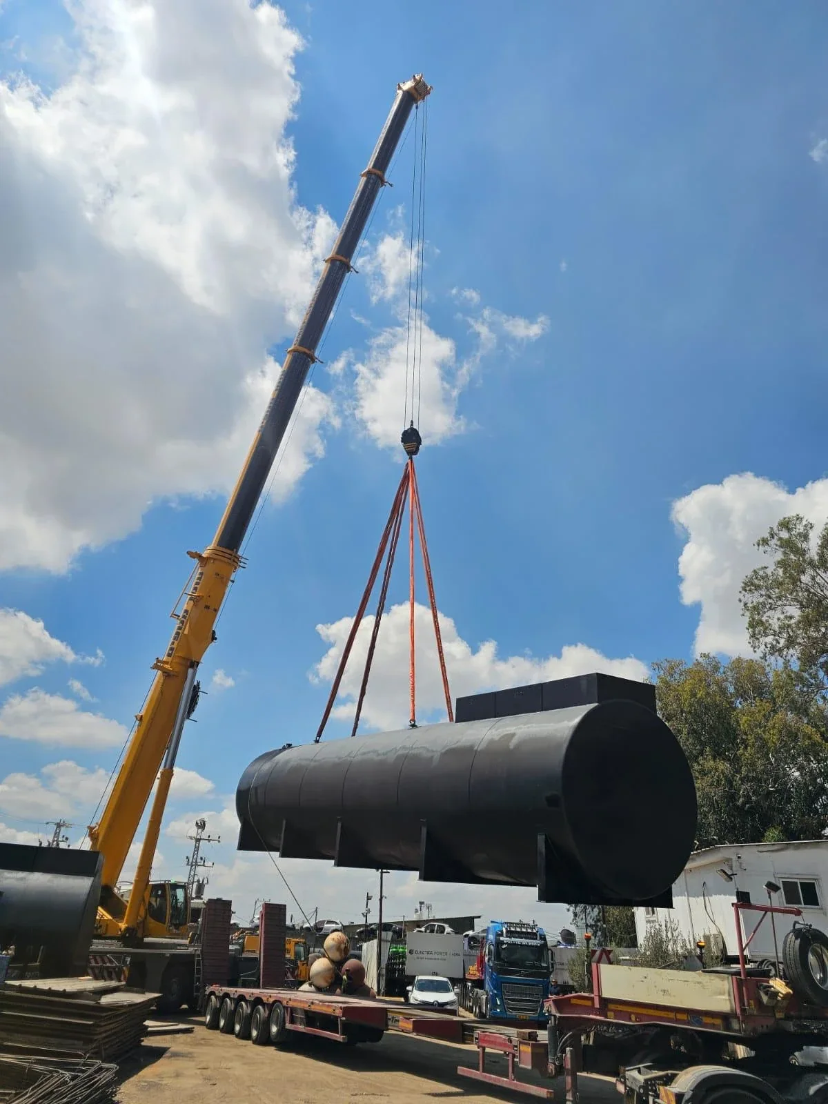 מנוף צהוב מניף מיכל דלק שחור ארוך ברתמות כתומות מעל מוביל, שמיים כחולים עם עננים