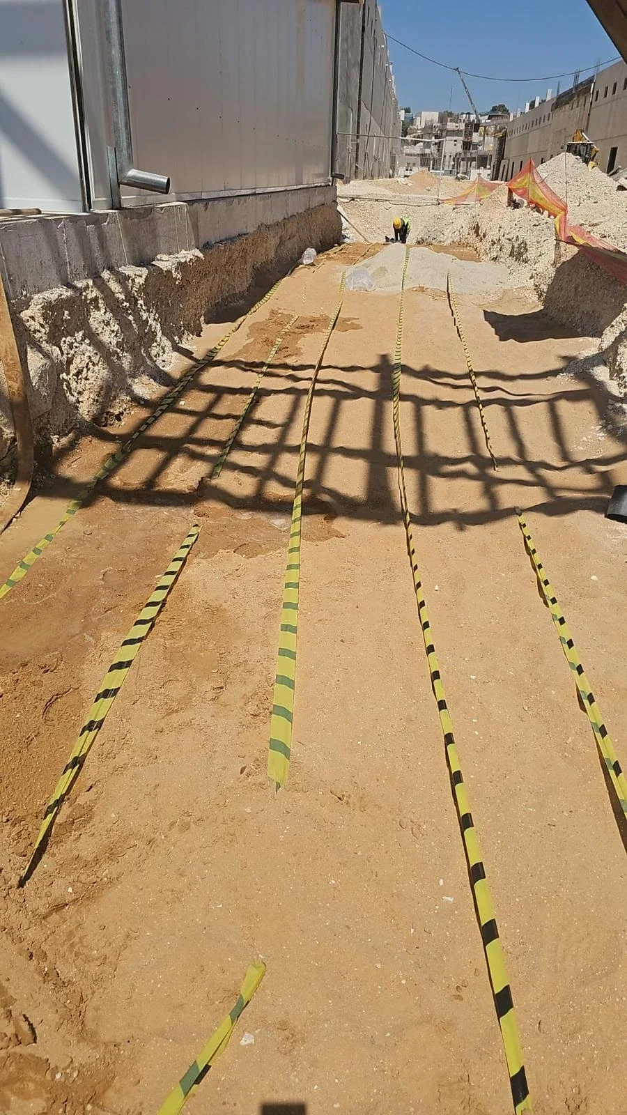 תעלה מכוסה חול עם סרטי סימון צהובים־שחורים לאורכה, מבנה בטון בצד ועובד ברקע