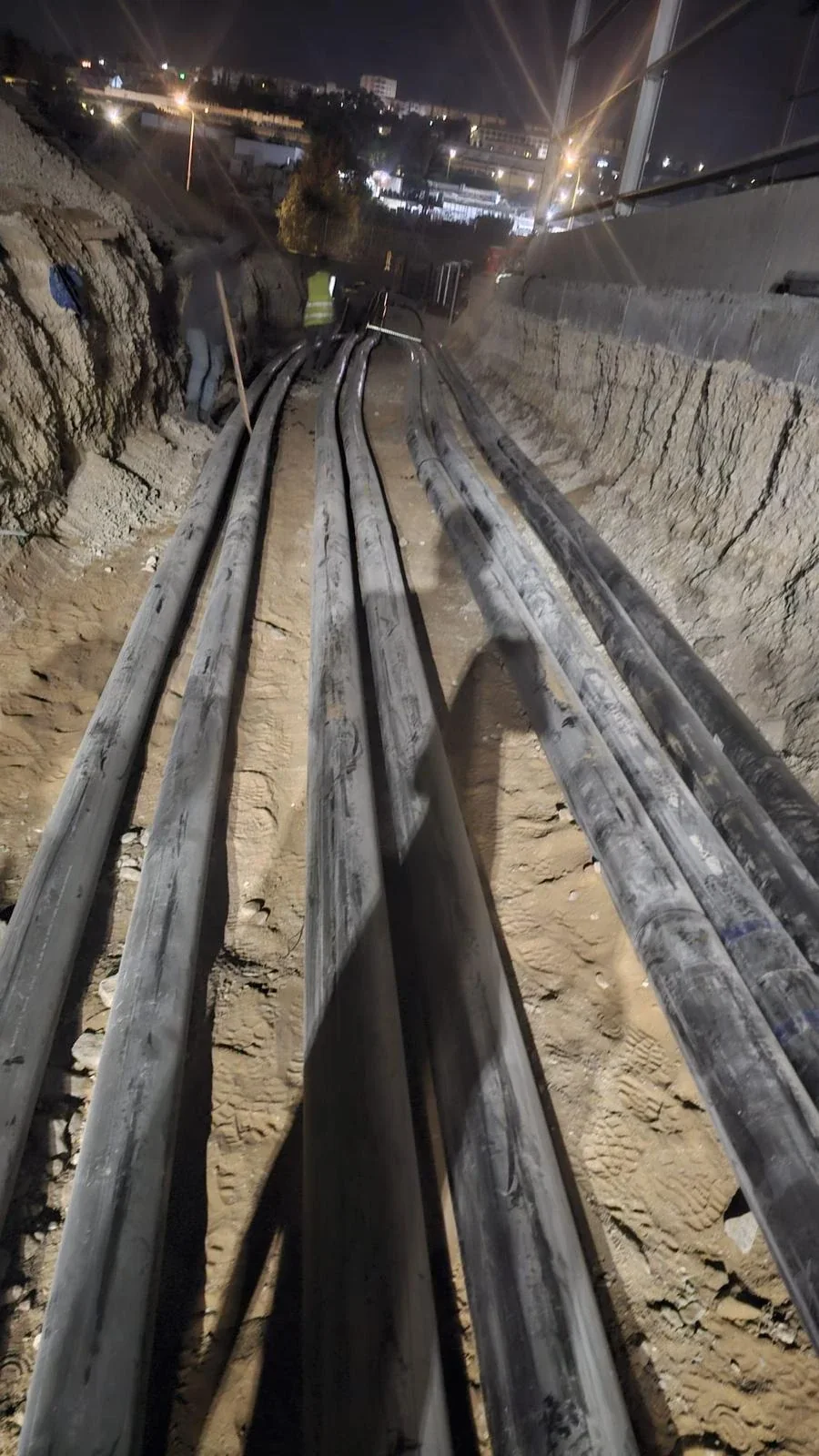 שמונה קווי צנרת שחורים מונחים בתעלה חפורה בלילה, עובד באפוד זוהר ברקע ואורות העיר