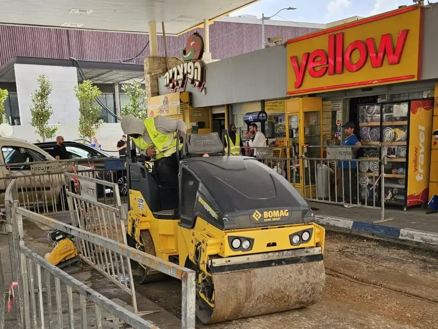 מכבש קטן מהדק את הרחבה בתחנת דלק פעילה, מאחורי גדר בטיחות