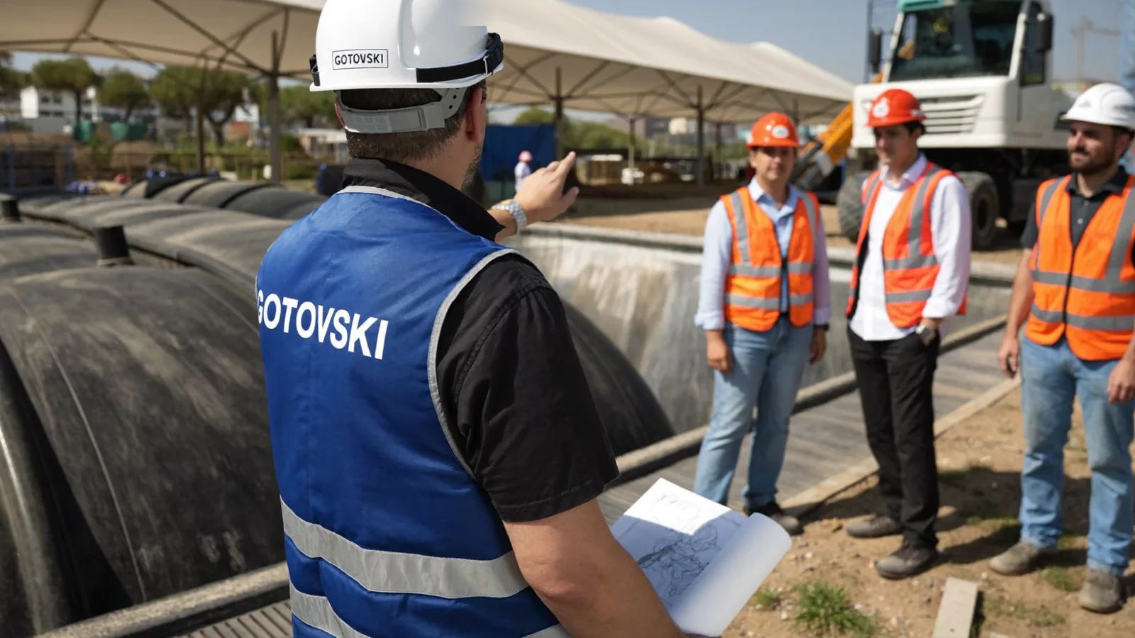 הדמיה: מנהל עבודה עם קסדה ואפוד גוטובסקי מדריך שלושה אנשי צוות ליד מיכלי דלק באתר תחנה בהקמה
